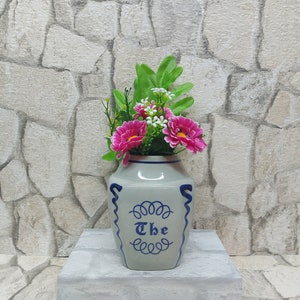 May include: A grey ceramic vase with a blue design and the word "The" in script. The vase is filled with pink and white artificial flowers and green leaves. The vase is on a grey surface.