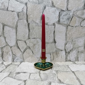 May include: A tall, red taper candle in a brass and green heart-shaped candle holder. The candle holder has gold star accents and a gold rim. The candle is standing on a stone surface.