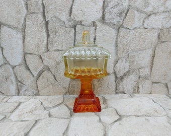 May include: A clear glass candy dish with a domed lid and a pedestal base. The base is orange and yellow.