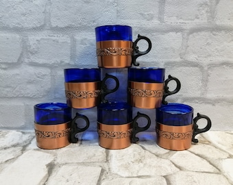May include: A set of six cobalt blue glass mugs, each with a copper-coloured metal filigree band and a black handle. The mugs are arranged in a pyramid shape. The background is a white brick wall.