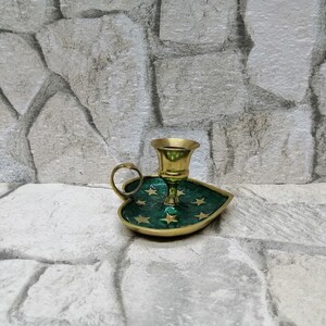 May include: A brass candle holder with a green leaf-shaped base adorned with gold stars. The candle holder has a small handle and a cup-shaped top to hold a candle. The background is a stone wall.