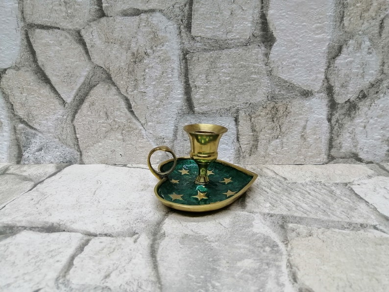 May include: A brass candle holder with a green leaf-shaped base adorned with gold stars. The candle holder has a small handle and a cup-shaped top to hold a candle. The background is a stone wall.