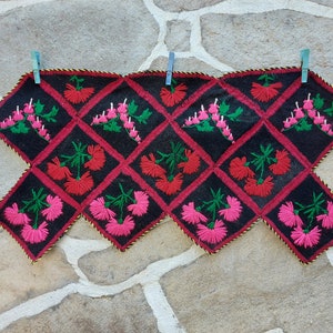 Könnte beinhalten: Schwarzes Textil mit Rautenmuster, roten Rändern und floralen Stickereien in Rosa, Rot und Grün. Das Textil ist mit goldenen Zierleisten versehen und wird von blauen Wäscheklammern gehalten. Ein dekoratives Stück.