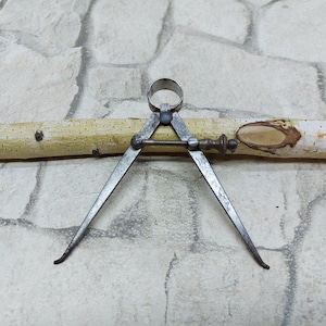 May include: An antique metal caliper tool with pointed legs, a central adjustment screw, and a ring at the top. The caliper is positioned over a light-colored wooden dowel with a natural bark pattern. The background is a textured stone surface.