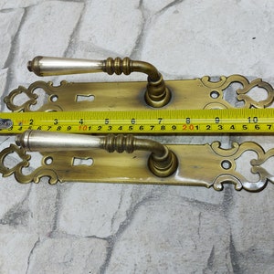 Op de afbeelding: Twee antieke messing deurklinken met sierlijke details. Elke klink heeft een zilverkleurige greep en een sleutelgat. De klinken zijn ongeveer 28 cm lang, gemeten met een gele rolmaat.