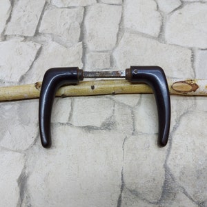 May include: Two dark brown door handles with a curved design are displayed. The handles are connected by a metal rod. The handles are resting on a light brown wooden stick, against a stone background.
