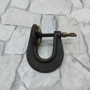 May include: A vintage C-clamp, a tool used to hold workpieces together. The clamp is made of dark metal with brass accents on the screw and handle. The U-shaped frame is sturdy, and the screw mechanism is visible.