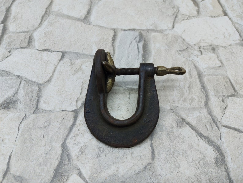 May include: A vintage C-clamp, a tool used to hold workpieces together. The clamp is made of dark metal with brass accents on the screw and handle. The U-shaped frame is sturdy, and the screw mechanism is visible.