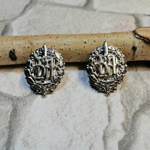 May include: Two silver-toned metal pins with a wreath design and the letters "DFH" in the center. The pins are on a wooden surface.