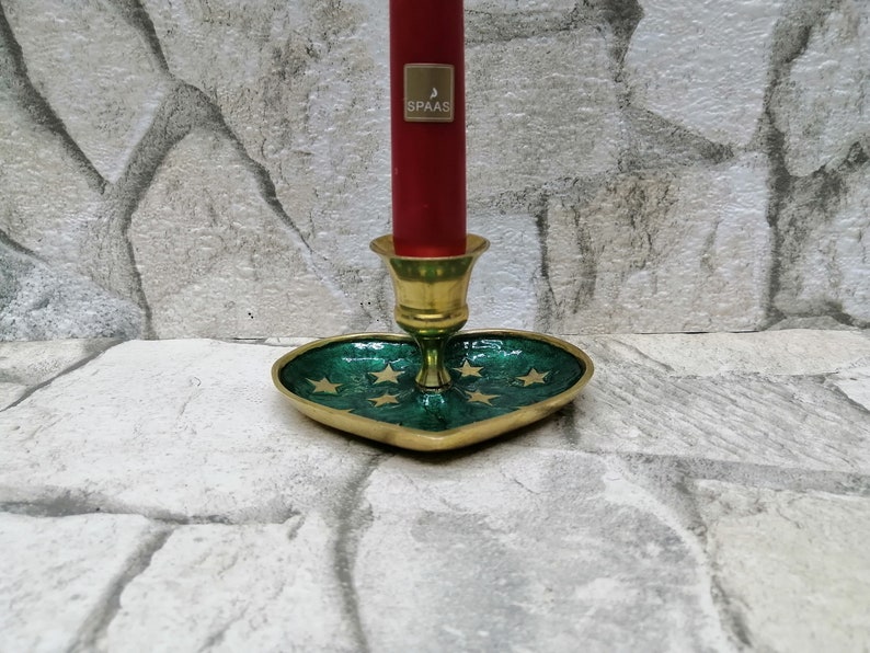 May include: A brass candle holder with a heart-shaped green base adorned with gold stars. A red candle with a gold label is inserted into the holder. The candle holder is a decorative item.