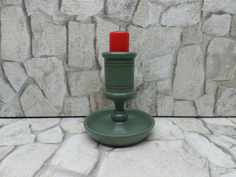 May include: A green wooden candlestick with a red candle. The candlestick has a round base and a detailed design. The candle is cylindrical and has a burnt wick. The background is a stone wall.