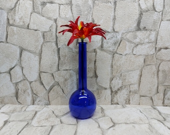 May include: A blue glass vase with a round base and a long, thin neck. A red and orange flower is in the vase.