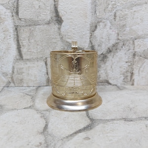 Könnte beinhalten: Ein goldfarbener Metall-Becherhalter mit einem verzierten Design, das ein Gebäude mit einem Springbrunnen zeigt. Der Becherhalter hat einen Griff und einen Sockel.
