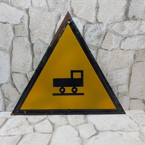 May include: A yellow triangular warning sign with a black border. The sign has a black silhouette of a truck with wheels on a track. The sign is on a stone wall background.