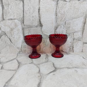 Könnte beinhalten: Zwei rubinrote Dessertschalen aus Glas mit gewellten Rändern. Die Schalen im Vintage-Stil haben einen Sockel und vertikale Rippendetails. Die Schalen stehen auf einer steinartigen Oberfläche.