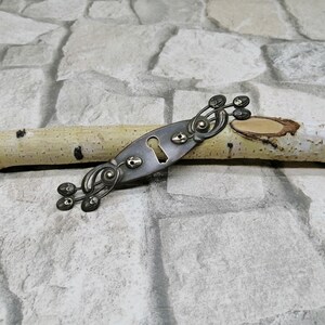 May include: An antique-style, silver-toned metal escutcheon plate with a keyhole and decorative flourishes. The plate is oval-shaped with ornate detailing and is resting on a light-colored birch branch against a stone background.