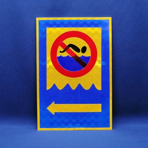 May include: A yellow and blue reflective sign with a red circle and a diagonal line through it. The circle contains a black silhouette of a person swimming. The sign also has a yellow arrow pointing to the left and the words "No Swimming" in black.