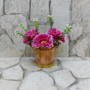 Peut inclure: Un petit seau en cuivre avec une base et une poignée en laiton, rempli de fleurs roses vives avec des bords blancs et des centres jaunes. Des brins verts et blancs ajoutent de la hauteur à l'arrangement floral. Le seau est posé sur une surface en pierre.