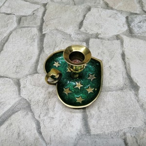 May include: A brass candle holder with a green enamel base shaped like a shield. The base is decorated with gold stars. The candle holder has a ring handle and a gold-colored candle cup.
