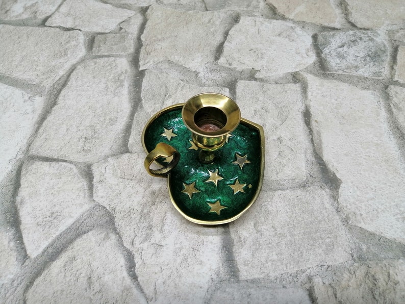 May include: A brass candle holder with a green enamel base shaped like a shield. The base is decorated with gold stars. The candle holder has a ring handle and a gold-colored candle cup.
