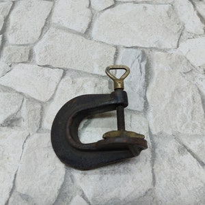 May include: A vintage C-clamp made of dark metal with brass accents. The clamp features a threaded rod for adjusting the opening and a brass handle. The tool is designed for holding objects together during woodworking or metalworking projects.