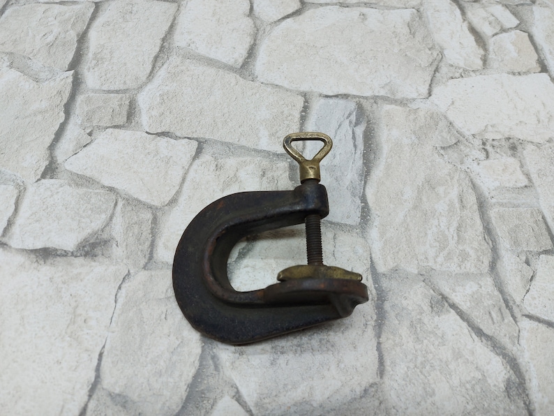 May include: A vintage C-clamp made of dark metal with brass accents. The clamp features a threaded rod for adjusting the opening and a brass handle. The tool is designed for holding objects together during woodworking or metalworking projects.