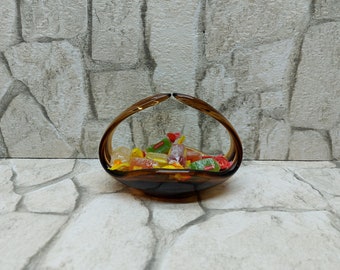 May include: A brown glass candy bowl with a handle, filled with colourful wrapped sweets. The bowl has a rounded base and a curved handle that rises from the sides. The sweets are various colours and shapes.