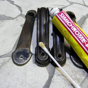 May include: An image shows four metal objects and a tube of Simichrome Polish. The metal objects are dark brown and have a curved shape. The tube of polish is yellow with red and white text. A cotton swab rests on one of the metal objects.
