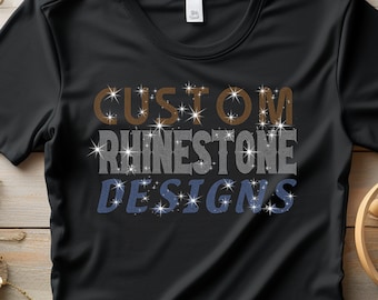 Op de afbeelding: Zwart t-shirt met de tekst "CUSTOM RHINESTONE DESIGNS" in bruin, grijs en blauw, elk woord versierd met sprankelende, stervormige strass-steentjes. Het shirt wordt gepresenteerd op een houten oppervlak met decoratieve elementen.