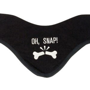 Può includere: Fascia per capelli nera con la scritta bianca "OH, SNAP!" e un'immagine di un osso rotto. La fascia ha una forma curva e un bordo nero.