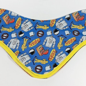 Può includere: Un bandana blu con un bordo giallo che presenta un motivo ripetuto di immagini a tema Seinfeld, tra cui il logo dello show, una tazza da caffè, una scatola di cereali e un uomo con una camicia bianca.
