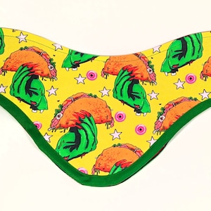 Può includere: Un bandana giallo con una mano di mostro verde che afferra un taco. Il bandana ha un bordo verde e rosso. Lo sfondo del bandana è giallo con occhi rosa e stelle bianche.