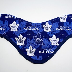 Op de afbeelding: Een blauwe stoffen hondenbandana met een wit Toronto Maple Leafs-logo dat zich herhaalt op de stof. De bandana is dubbelgevouwen en de randen zijn gestikt.