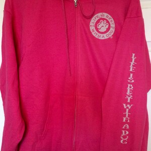 May include: A bright pink zip-up hoodie with a drawstring hood. The left sleeve has the text "LIFE IS BEST WITH A DOG" printed vertically. The chest features a circular graphic with a paw print and the same text.