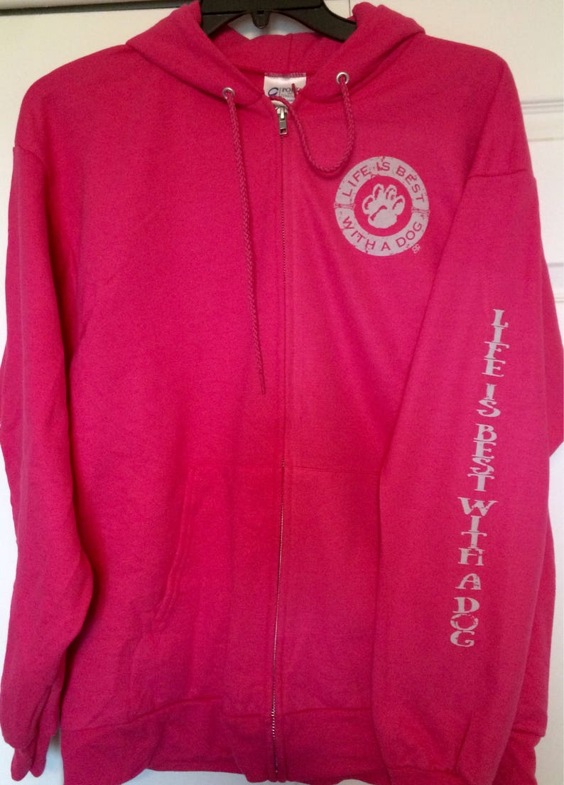 May include: A bright pink zip-up hoodie with a drawstring hood. The left sleeve has the text "LIFE IS BEST WITH A DOG" printed vertically. The chest features a circular graphic with a paw print and the same text.