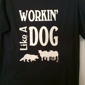 May include: Black t-shirt with the phrase "Workin' Like A Dog" in distressed white lettering. A dog silhouette and sheep silhouettes are below the text, suggesting a work theme.