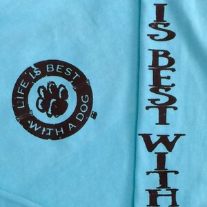 May include: Light blue long-sleeve shirt with the text "LIFE IS BEST WITH A DOG" printed in brown. The phrase is printed vertically on the sleeve and in a circular design on the chest, featuring a paw print.