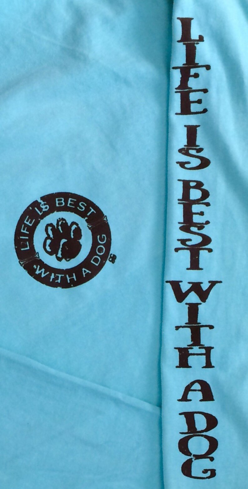 May include: Light blue long-sleeve shirt with the text "LIFE IS BEST WITH A DOG" printed in brown. The phrase is printed vertically on the sleeve and in a circular design on the chest, featuring a paw print.