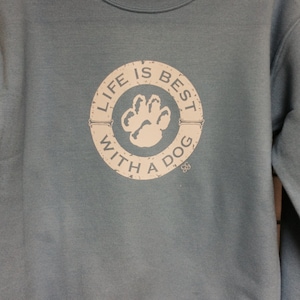 May include: A light blue sweatshirt with a crew neck. The front features a circular graphic with the text "LIFE IS BEST WITH A DOG" surrounding a white paw print. The graphic has a distressed, vintage look.