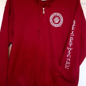 May include: A red zip-up hoodie with a hood and front pockets. The left sleeve has the text "LIFE IS BEST WITH A DOG" printed vertically. The chest features a circular design with a paw print and the same text.