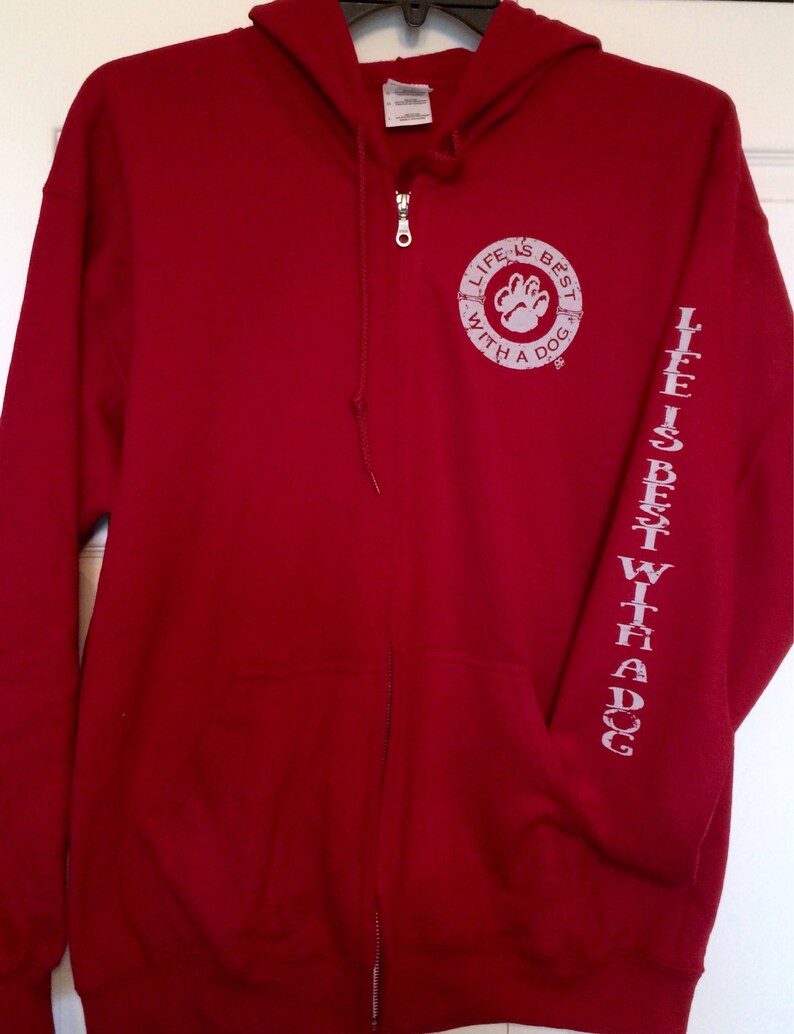 May include: A red zip-up hoodie with a hood and front pockets. The left sleeve has the text "LIFE IS BEST WITH A DOG" printed vertically. The chest features a circular design with a paw print and the same text.