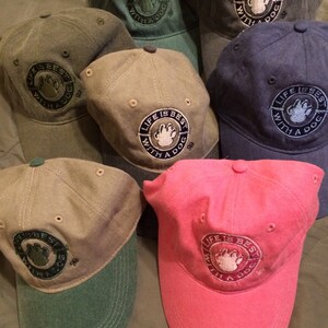 May include: A collection of baseball caps in various colors, including pink, blue, and green. Each cap features an embroidered circular patch with the text "LIFE IS BEST WITH A DOG" and a paw print.