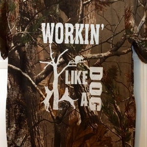 May include: Camouflage pattern t-shirt with the text "WORKIN' LIKE A DOG" in white block letters. The shirt features a crew neck and short sleeves. The design includes a tree and dog silhouettes.