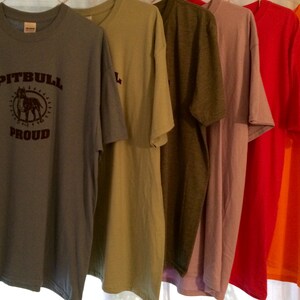 May include: A row of six short-sleeved t-shirts in various colors, including blue-grey, olive green, mauve, red, and orange. The first shirt has the text "PITBULL PROUD" with a graphic of a dog. The shirts are hanging on a white rod.