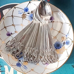 Può includere: Un set di cucchiaini d'argento con manici decorati sono impilati su un piattino bianco con un motivo floreale. Il piattino è su una superficie blu con le parole "Eastern League New York 19 Champ" stampate su di esso.