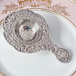 Peut inclure: Une passoire à thé en argent avec un motif floral orné repose sur une assiette rose et blanche avec des accents dorés. L'assiette a un bord festonné et des motifs floraux. La passoire est détaillée avec des gravures complexes.