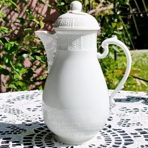 Pode incluir: Uma cafeteira de porcelana branca com tampa e alça, colocada sobre uma toalha de mesa de renda branca. A cafeteira tem um corpo arredondado e uma tampa texturizada. O fundo apresenta folhagem verde e luz solar.
