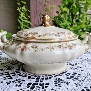 Può includere: Piatto da portata color crema con coperchio, motivi floreali e dettagli dorati. Il piatto ha due manici e un pomello decorativo sul coperchio. Il motivo floreale comprende fiori rosa, gialli e blu. Il piatto è appoggiato su una tovaglia di pizzo bianco.