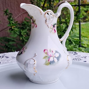 May include: A white porcelain jug with a curved spout and handle, decorated with pink and white floral patterns and gold accents. The jug is on a white tray with a lace doily.