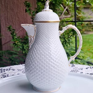 May include: White ceramic coffee pot with a textured diamond pattern, gold trim, and a matching lid. The pot has a curved handle and is sitting on a white oval tray with gold accents. The background includes greenery and a white lace tablecloth.